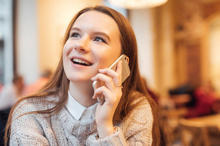 快乐的女孩坐在咖啡馆里，用快乐的脸在电话里交谈。 穿白色毛衣。 冬季秋装。