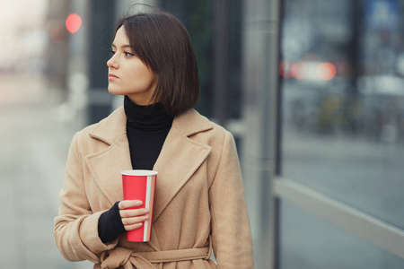 女人在街上散步时喝她的热咖啡。 穿着冬天衣服喝热咖啡的时尚微笑女人的肖像。 女性冬季风格。 图像