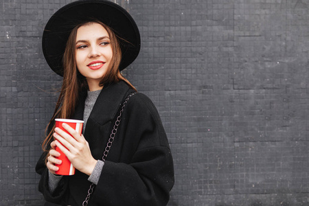 年轻的时尚女性拿着她的热咖啡杯站在墙附近，戴着黑色帽子环顾四周，微笑着对着镜头。 复制空间。 图像