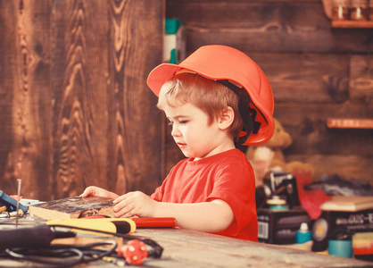 忙碌的脸上的幼儿在车间附近的桌子上玩工具。车间和工具概念。孩子在头盔, 硬帽可爱的发挥作为建设者或修理工, 手工制作。玩乐器的孩