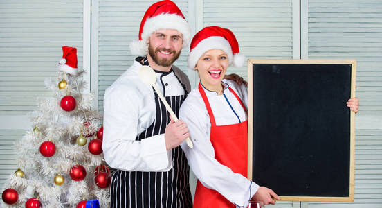 男女厨师圣塔帽子在圣诞树附近举行黑板复制空间。夫妇检查圣诞晚餐的清单成分。烹饪圣诞大餐。圣诞食谱的概念。我们家庭的菜单