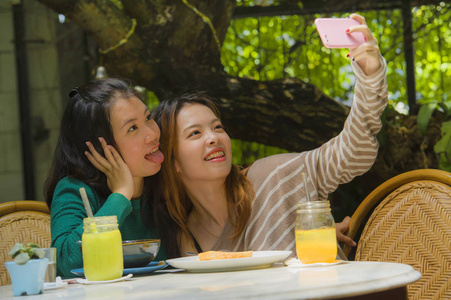 两位年轻快乐可爱的亚洲女朋友用手机相机拍摄自拍肖像画，用于网络社交媒体，同时在咖啡馆吃健康早午餐