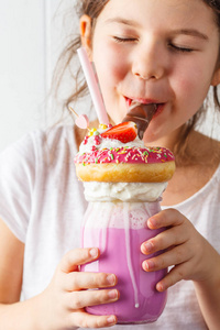 小微笑的女孩喝粉红色草莓怪物奶昔鸡尾酒与甜甜圈和糖果白色背景。 不健康的甜点概念。