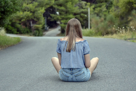 穿牛仔服的旅游女孩坐在树林里的路上