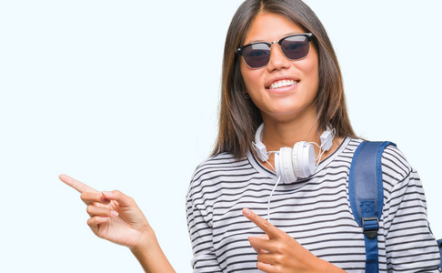 年轻的亚洲学生妇女戴着耳机和背包在孤立的背景上微笑着，看着相机，用两只手和手指指向一边。
