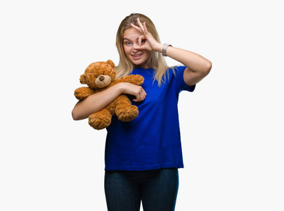 年轻的白种人女人拿着可爱的泰迪熊，在孤立的背景上，快乐的脸，微笑着，做着好的标志，用手看着手指。