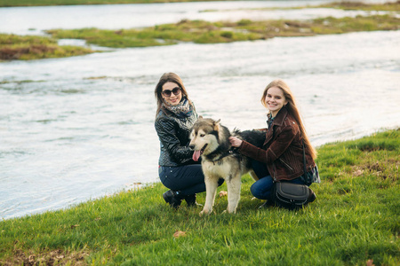 两姐妹带着哈士奇狗走在河前。布鲁内特和金发碧眼的女孩爱抚狗