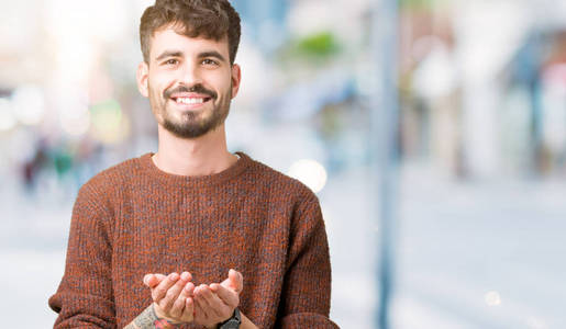 年轻的英俊男子穿着冬天的毛衣，在孤立的背景上微笑，双手一起接受或给予手势。 持有和保护
