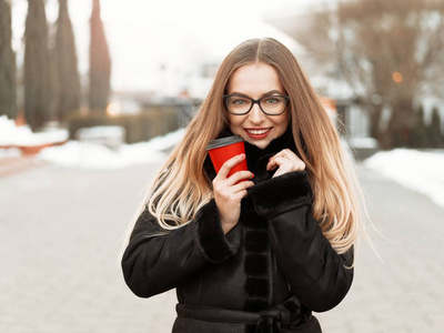 年轻美丽开朗的女孩走过冬天的城市，从红色的玻璃杯里喝一杯