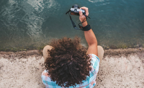 从上面看到的年轻男性卷发检查照片的自然在他的数码相机。 英俊的男人穿着休闲衬衫，在湖边摆着数码相机。 人们旅行生活方式