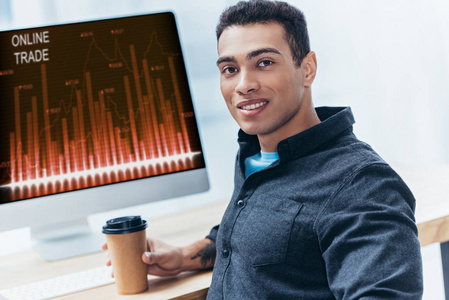 年轻的商人带着咖啡，带着台式电脑和在线贸易图表，对着相机微笑