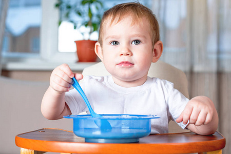 小男孩学着用勺子吃饭，他自己在厨房的儿童餐桌上。健康婴儿食品的概念