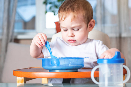 小男孩学着用勺子吃饭，他自己在厨房里的桌子上。 健康婴儿食品的概念