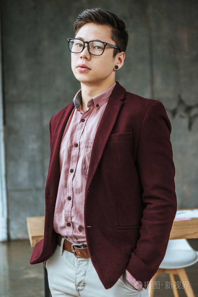 戴眼镜的年轻帅哥双手插着口袋在办公室里看着相机