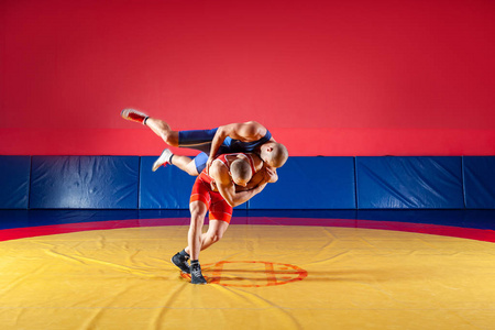 两个穿着蓝色和红色摔跤紧身衣的年轻人正在健身房里摔跤，在黄色的摔跤地毯上做一个臀部投掷。公平摔跤的概念