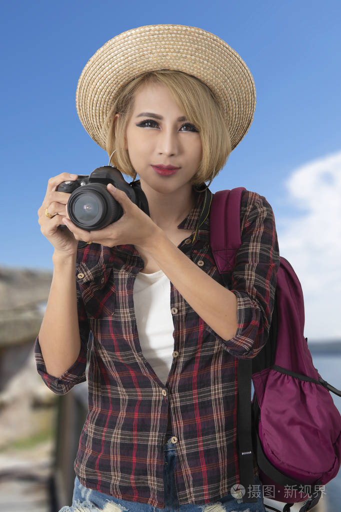 半拍照片的旅游女孩穿着格子衬衫,手中拿着相机,背景上有明亮的蓝天.