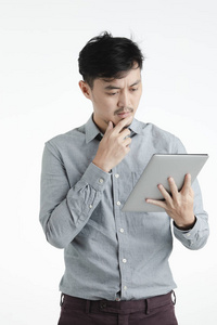 半长画室的黑发肖像，穿着灰色衬衫的亚洲年轻人，拿着数字平板电脑，盯着数字平板电脑，怀疑，困惑的样子，皱眉的脸，手放在下巴上，在孤