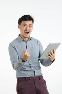 半长画室的黑发肖像，穿着灰色衬衫的亚洲年轻人，紧握拳头，张开嘴，满意地摆出一副姿势，拿着平板电脑，直视着在孤立的白色背景上。