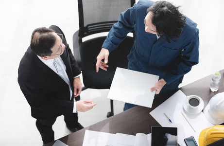 亚洲商人和男领班的俯视图,手里拿着纸,说话,忙着用黄白色安全帽