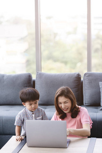 亚洲家庭幸福的时光，年轻漂亮的母亲，坐在地板上，可爱的蹒跚学步的孩子，在她的大腿上，深情地为他指着笔记本屏幕上的沙发背景
