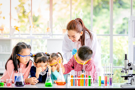 科学老师在实验室教亚洲学生，男孩把一些东西扔进橙色管，彩色试管和显微镜上的桌子概念，以便在实验室学习。