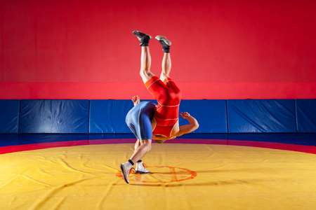 两个穿着蓝色和红色摔跤紧身衣的年轻人正在健身房里的黄色摔跤地毯上摔跤，并做了一复杂的摔跤。 公平摔跤的概念