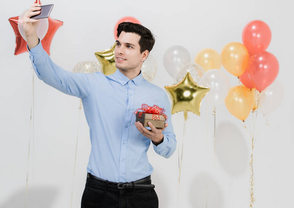 好看的年轻白种人男子拿着一个礼品盒，快乐的自拍在有趣的聚会白色背景与节日的彩色气球。