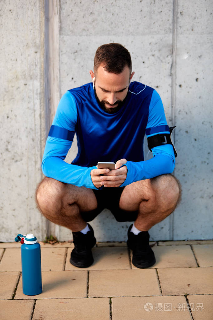 小胡子蹲着用智能手机. 在他旁边的瓶子里有水. 在背景墙上.