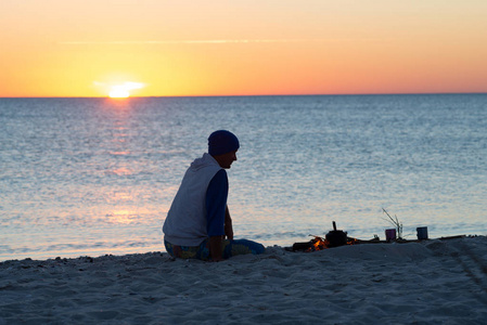 旅行者坐在海边的篝火旁沉思，欣赏海上的日落。 去海边旅行真棒。