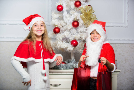 圣诞快乐。男孩孩子打扮成圣诞老人与白色人造胡子和红色帽子给礼物盒给女孩。孩子们用礼物庆祝圣诞节。圣诞老人带了一些礼物。打开圣诞礼