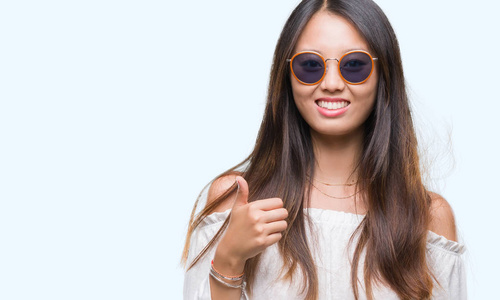 年轻的亚洲女人戴着太阳镜，在孤立的背景上，用手做快乐的竖起大拇指的手势。 赞许的表情看着相机，显示出成功。