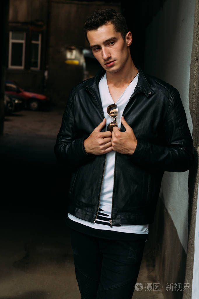 年轻的帅哥穿着白色t恤,黑色皮夹克和黑色裤子在街上戴着t恤太阳镜.