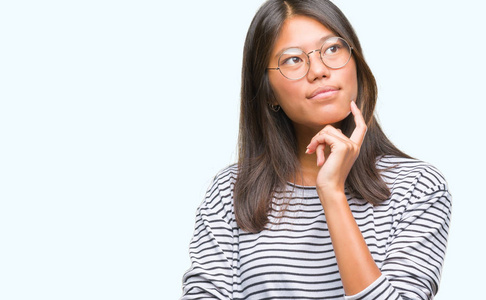 年轻的亚洲女人戴着眼镜，戴着孤立的背景，手拿着下巴，想着问题的表情。 带着体贴的脸微笑。 怀疑概念。