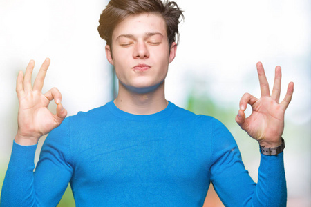 年轻的帅哥穿着蓝色毛衣，在孤立的背景上放松，微笑着，闭着眼睛，用手指做冥想手势。 瑜伽概念。