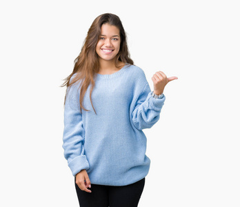 年轻漂亮的黑发女人穿着蓝色的冬季毛衣，在孤立的背景上微笑，快乐的脸看着，用拇指指向一边。