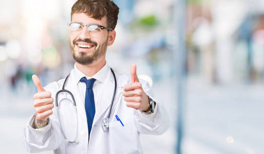 年轻的医生穿着医院的外套，在孤立的背景上，批准用手竖起大拇指做积极的手势，微笑着，为成功而高兴。 看着相机赢家的手势。