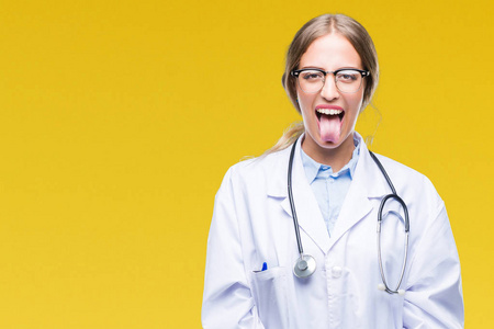 美丽的年轻金发医生女人穿着医疗制服，在孤立的背景下伸出舌头，以有趣的表情高兴。 情感概念。