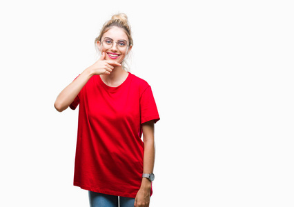 年轻漂亮的金发女人穿着红色的T恤和眼镜，戴着孤立的背景，自信地看着相机，微笑着交叉的手臂和举起的手在下巴上。 积极思考。