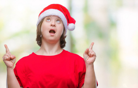 年轻的成年妇女戴着圣诞帽，戴着孤立的背景，惊讶地抬头，用手指和举起的手臂指着。