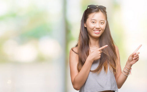 年轻的亚洲女人戴着太阳镜在孤立的背景上微笑着，看着相机，用两只手和手指指向一边。