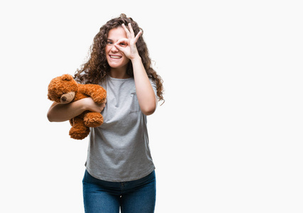 年轻的黑发女孩抱着玩具熊在孤立的背景上，快乐的脸，微笑着，做着好的标志，用手盯着手指看