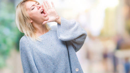 年轻漂亮的金发女人穿着冬天的毛衣，在孤立的背景上大声喊叫和尖叫，手在嘴上。 交流概念。