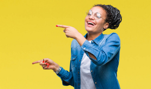 年轻的辫子非洲裔美国女孩戴着眼镜，在孤立的背景上微笑，看着相机，用两只手和手指指向一边。