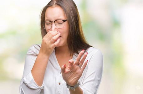 年轻的白种人漂亮的商业女人戴着眼镜，在孤立的背景上闻着一些臭味和令人恶心的难以忍受的气味，用手指在鼻子上屏住呼吸。 难闻的概念。