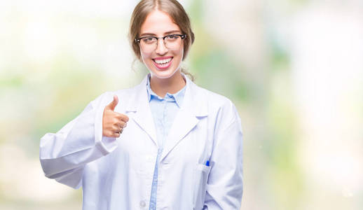 美丽的年轻金发医生女人穿着白色外套，在孤立的背景下，用手做快乐的竖起大拇指的手势。 赞许的表情看着相机，显示出成功。