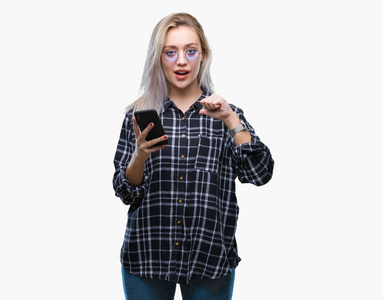 年轻的金发女人用智能手机在孤立的背景上发短信，惊讶的脸指向自己