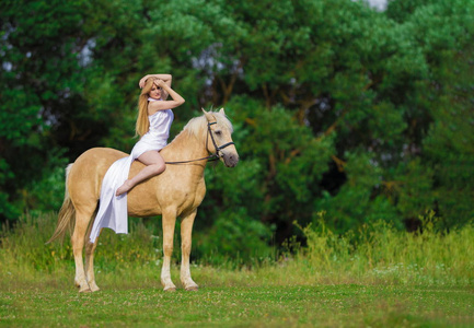 一位年轻的骑手,金发碧眼,身穿白色连衣裙,在田野和森林背景下骑着