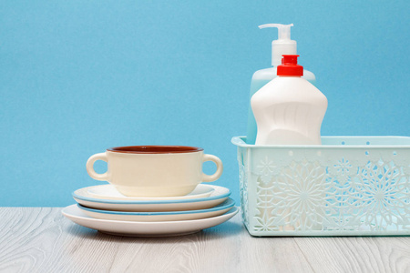 塑料瓶的洗碗液，玻璃和瓷砖清洁剂在篮子，干净的盘子和碗的蓝色背景。 洗涤和清洁的概念。