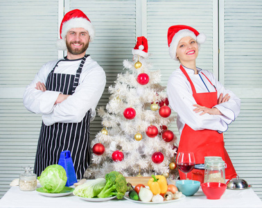 夫妇一起为圣诞晚餐准备健康的晚餐。男人和女人厨师围裙圣诞老人帽子附近的圣诞树。圣诞食谱的概念。烹饪圣诞大餐。秘密的成分是爱
