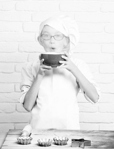 白色的制服的年轻男孩小可爱厨师厨师和帽子上有趣的脸，戴着眼镜站附近与轧制表针模具的蛋糕拿着红色的碗和吹进面粉对砖的背景墙上